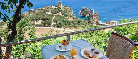 Una mesa servida con alimentos para el desayuno, incluyendo fruta, pastelería y una jarra de jugo, con vista a un paisaje costero con ruinas y vegetación.
