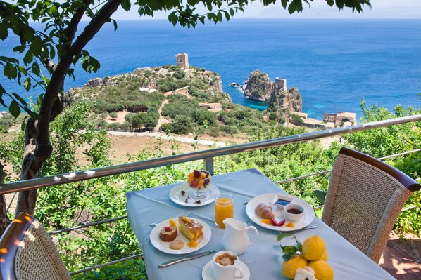 A table set with breakfast items, including fruit, pastries, and a jug of juice, overlooking a coastal landscape with ruins and greenery.