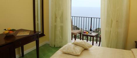 Un dormitorio con cama, mesita de noche, espejo y vista al mar a través de cortinas translúcidas.