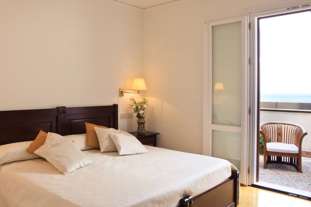 A hotel room with a bed, a nightstand with a lamp, a vase with flowers, and a view of the outside balcony.