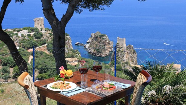 Una mesa con comida y bebidas, con vistas a un paisaje costero que incluye una torre y formaciones rocosas.