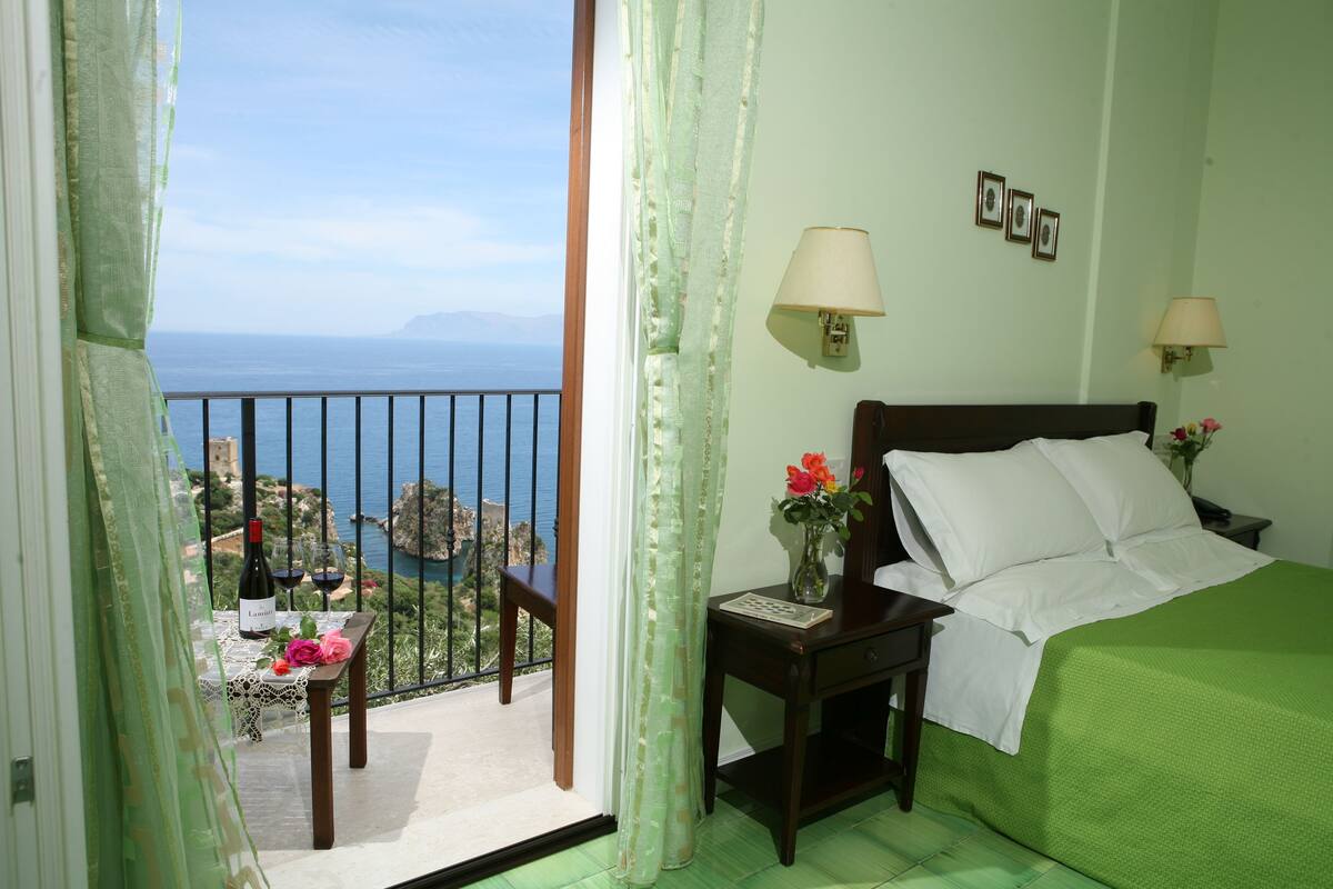 A hotel room with a balcony, a bed with white linens, a nightstand with a lamp, and a view of the sea.