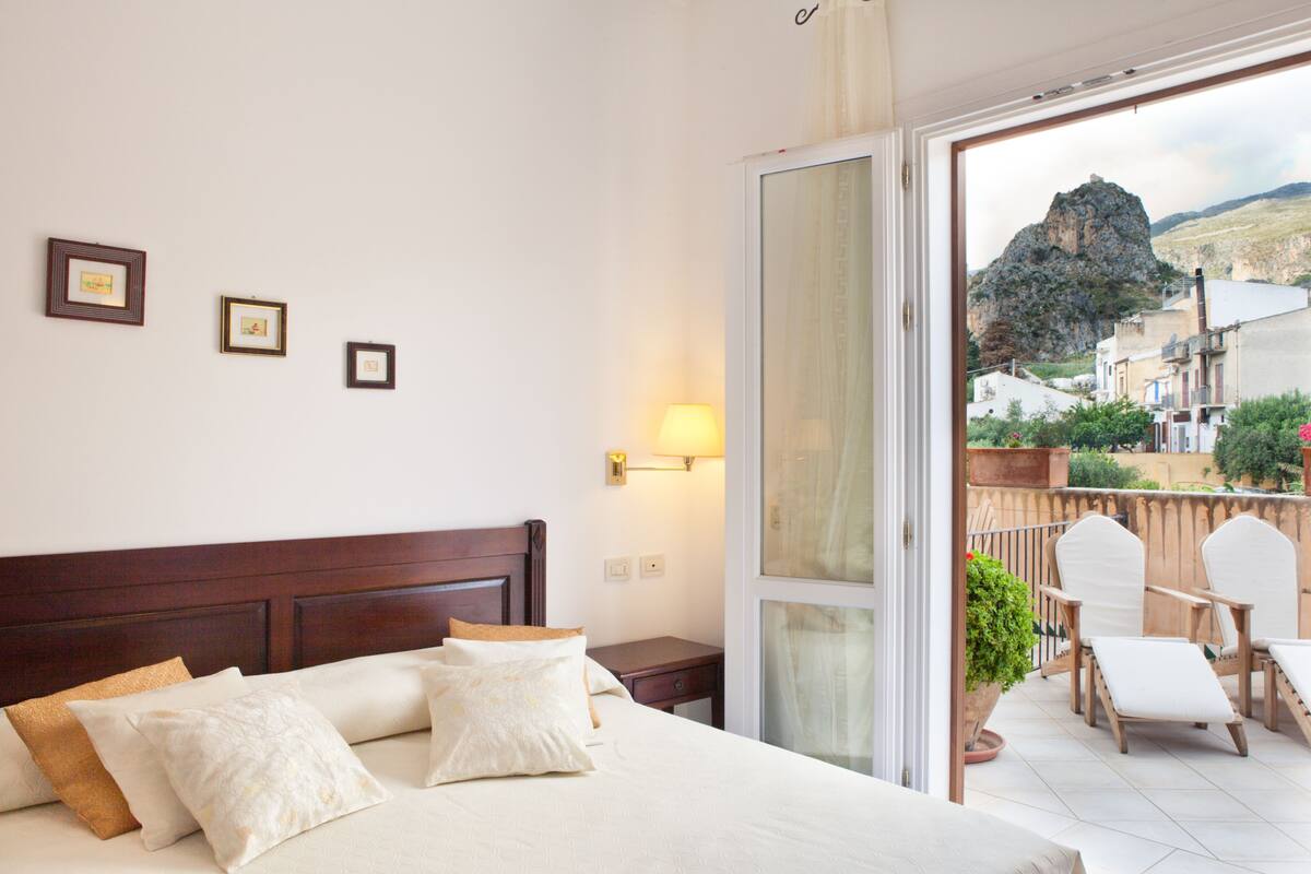A hotel room with a bed, pillows, a nightstand, a lamp, and a view of a balcony with chairs and a potted plant.