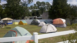 Un camping con varias tiendas de distintos tamaños y colores, una cerca blanca de piquetes y árboles al fondo.