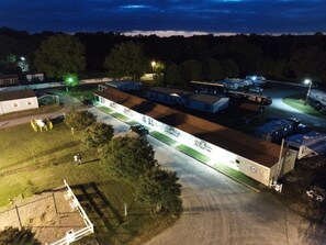 Um amplo edifício industrial com várias seções, cercado por veículos estacionados e algumas pessoas.