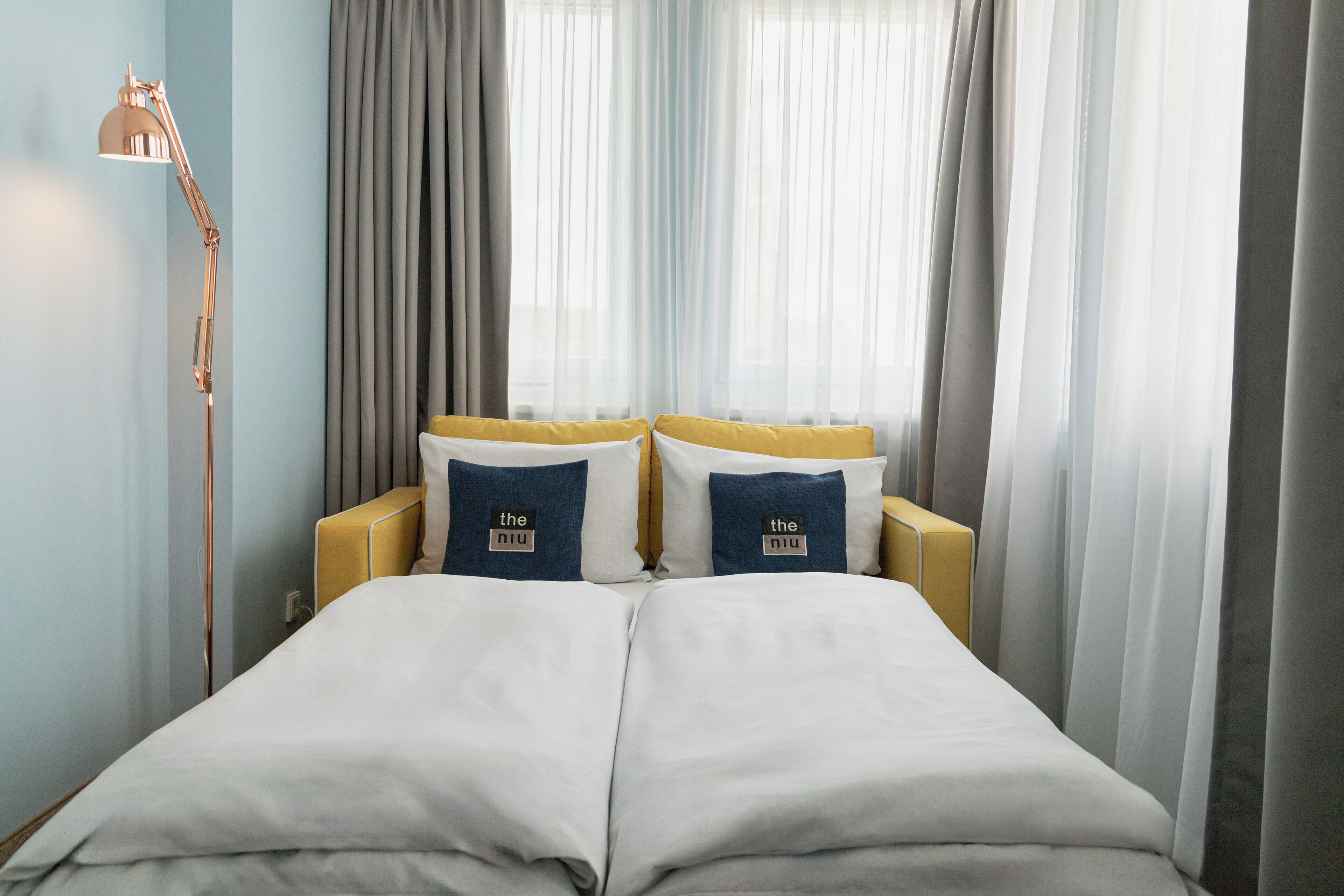 Una cama doble con dos almohadas, una lámpara de noche y cortinas traslúcidas.