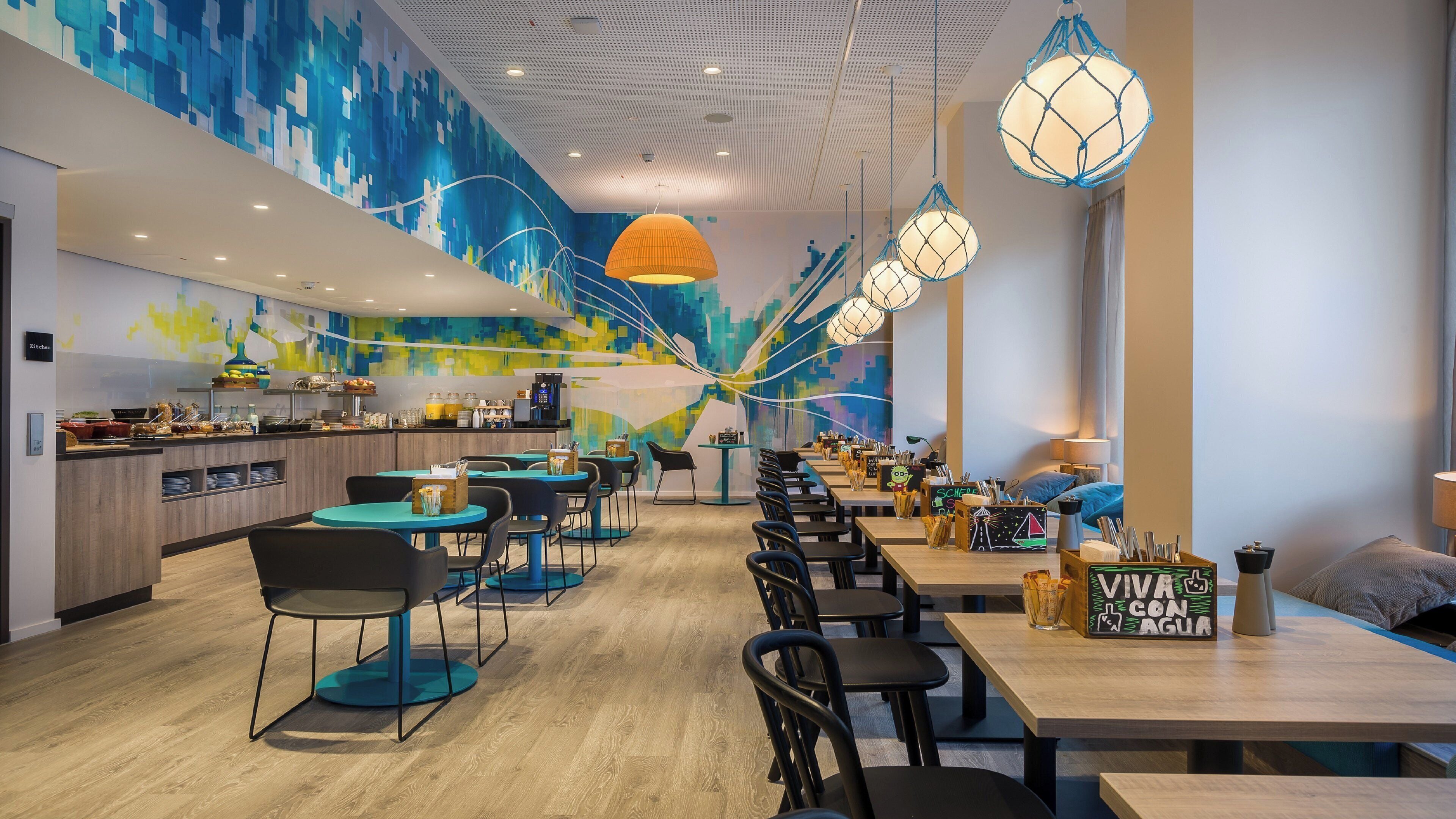 A modern restaurant with a long counter, multiple seating areas, and a colorful abstract mural on the wall.