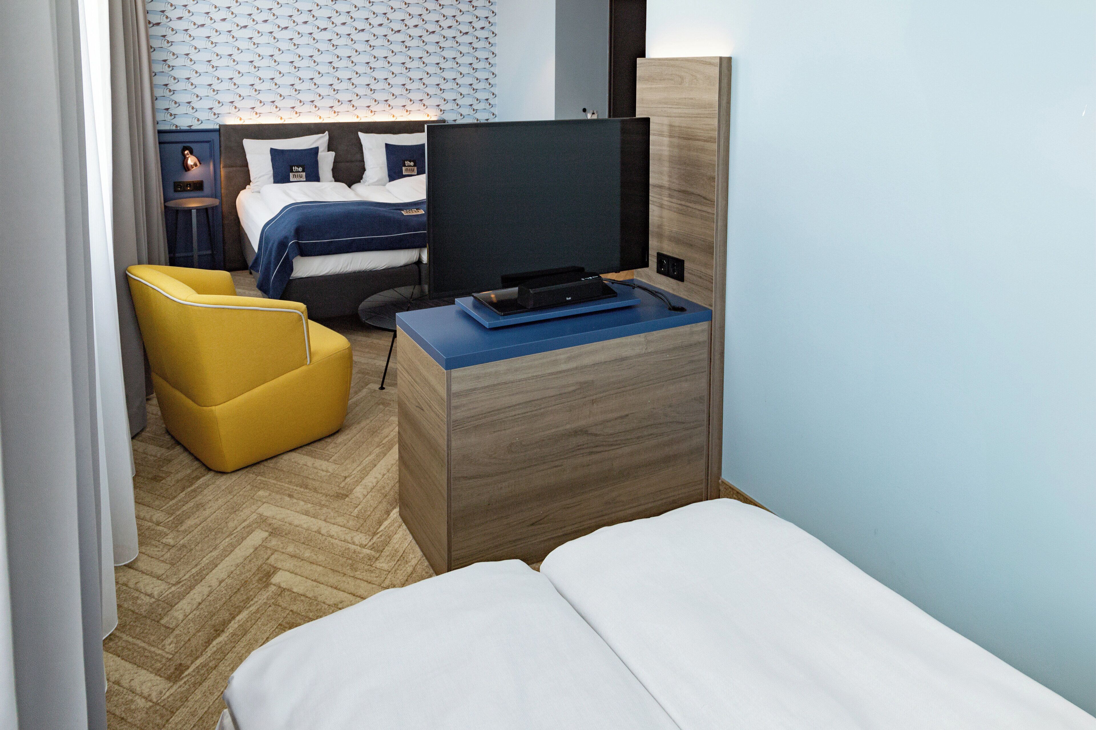 A hotel room with a bed, a television on a wooden dresser, a yellow armchair, and a small table.
