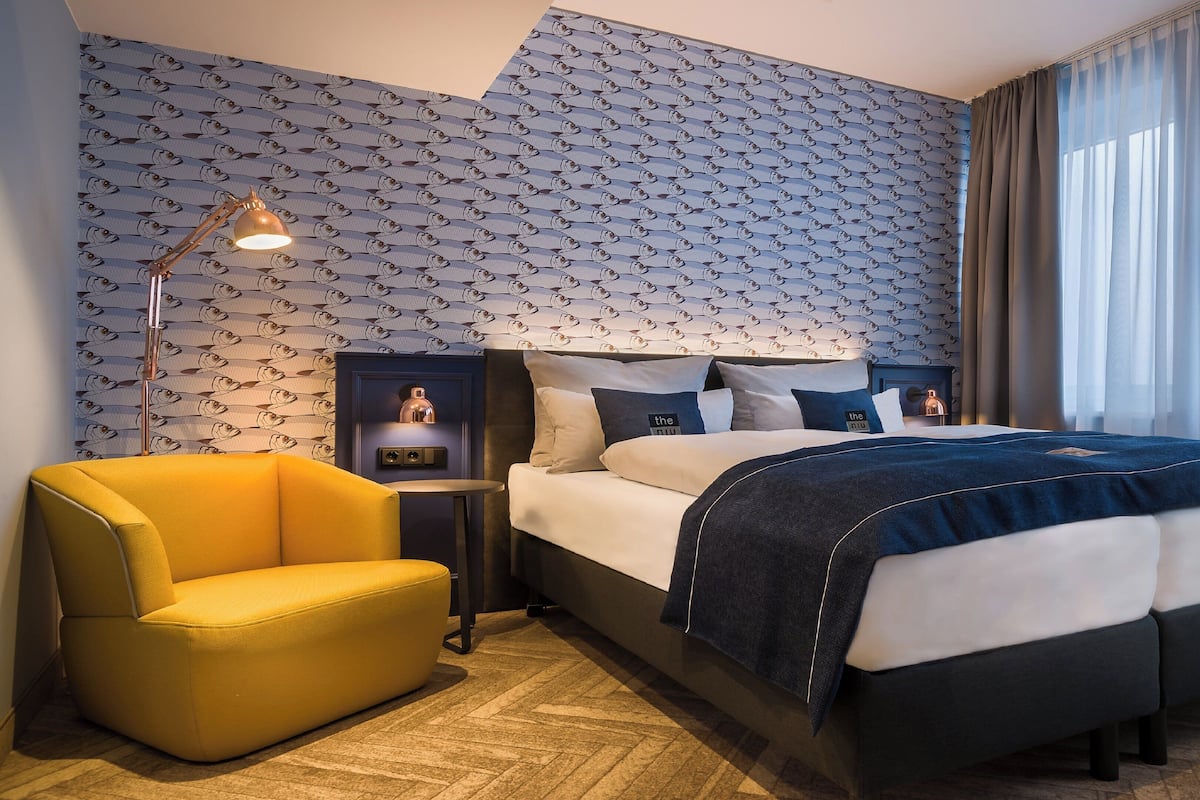 A hotel room with a bed, a desk, a chair, a lamp, and a patterned wallpaper.