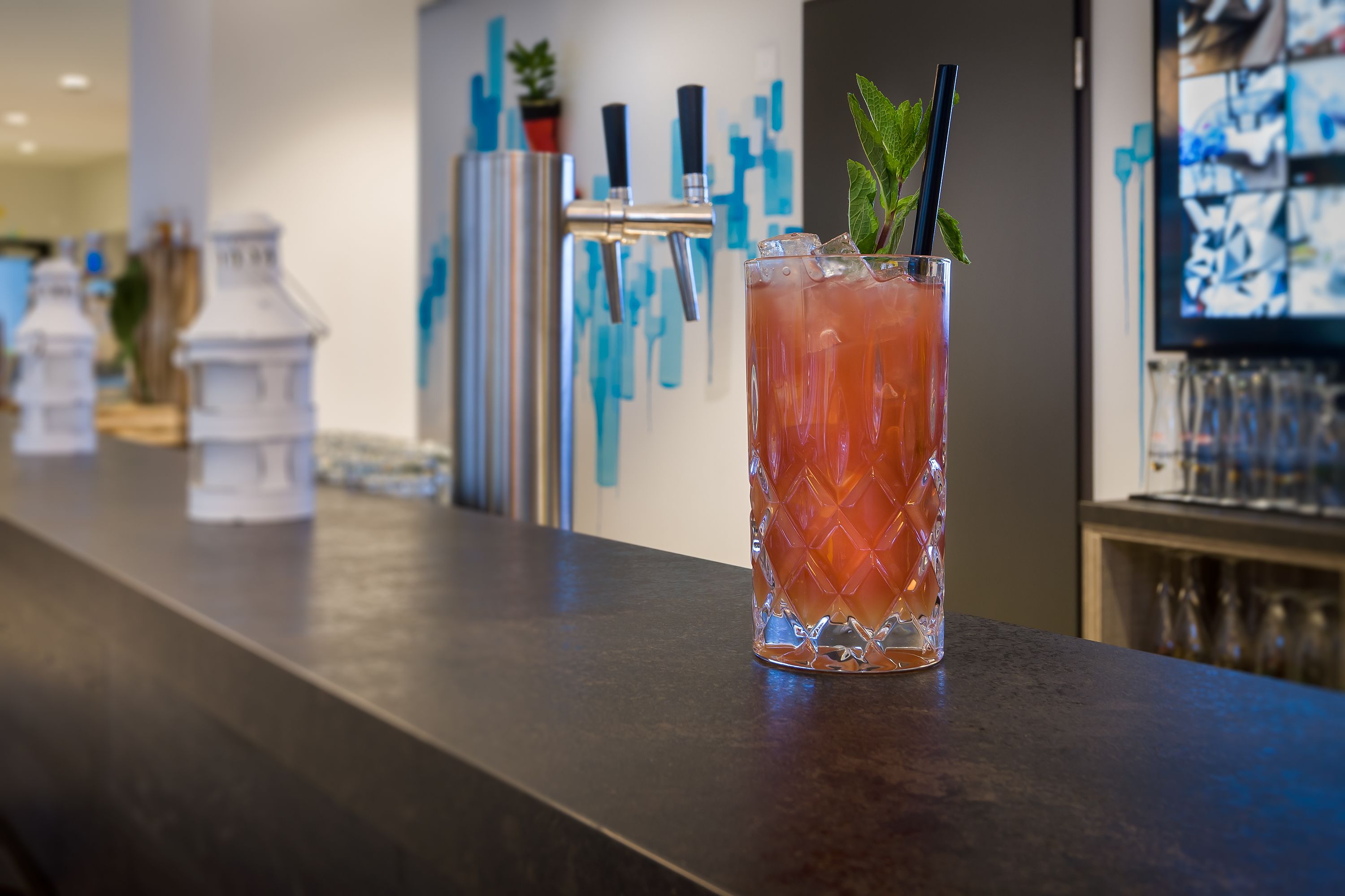 A bar area with a tall glass of a pink beverage, a faucet, and a decorative wall.