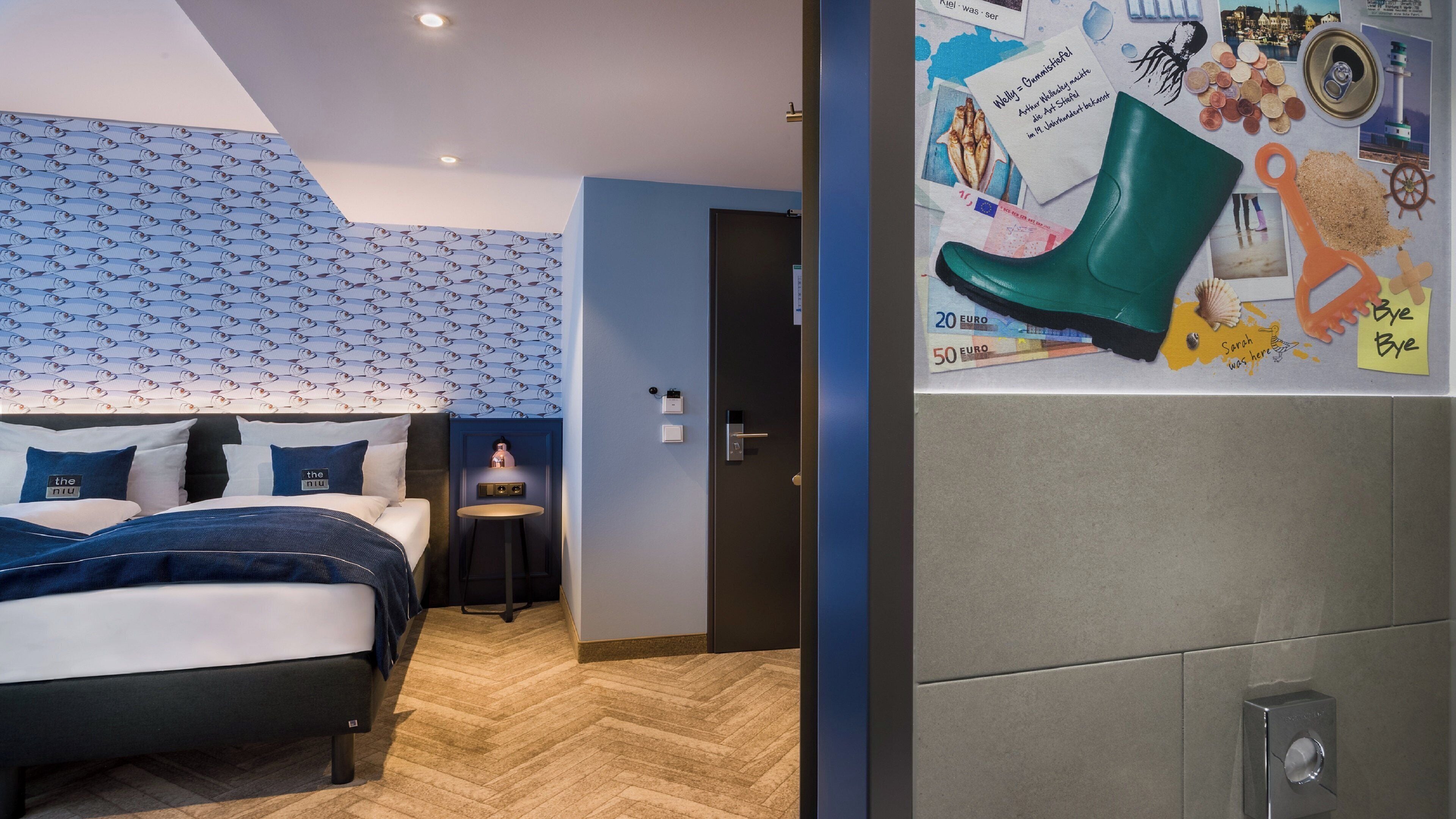 A hotel room with a bed, a small table, and a wall decorated with various items including a green boot, coins, and a lighthouse picture.