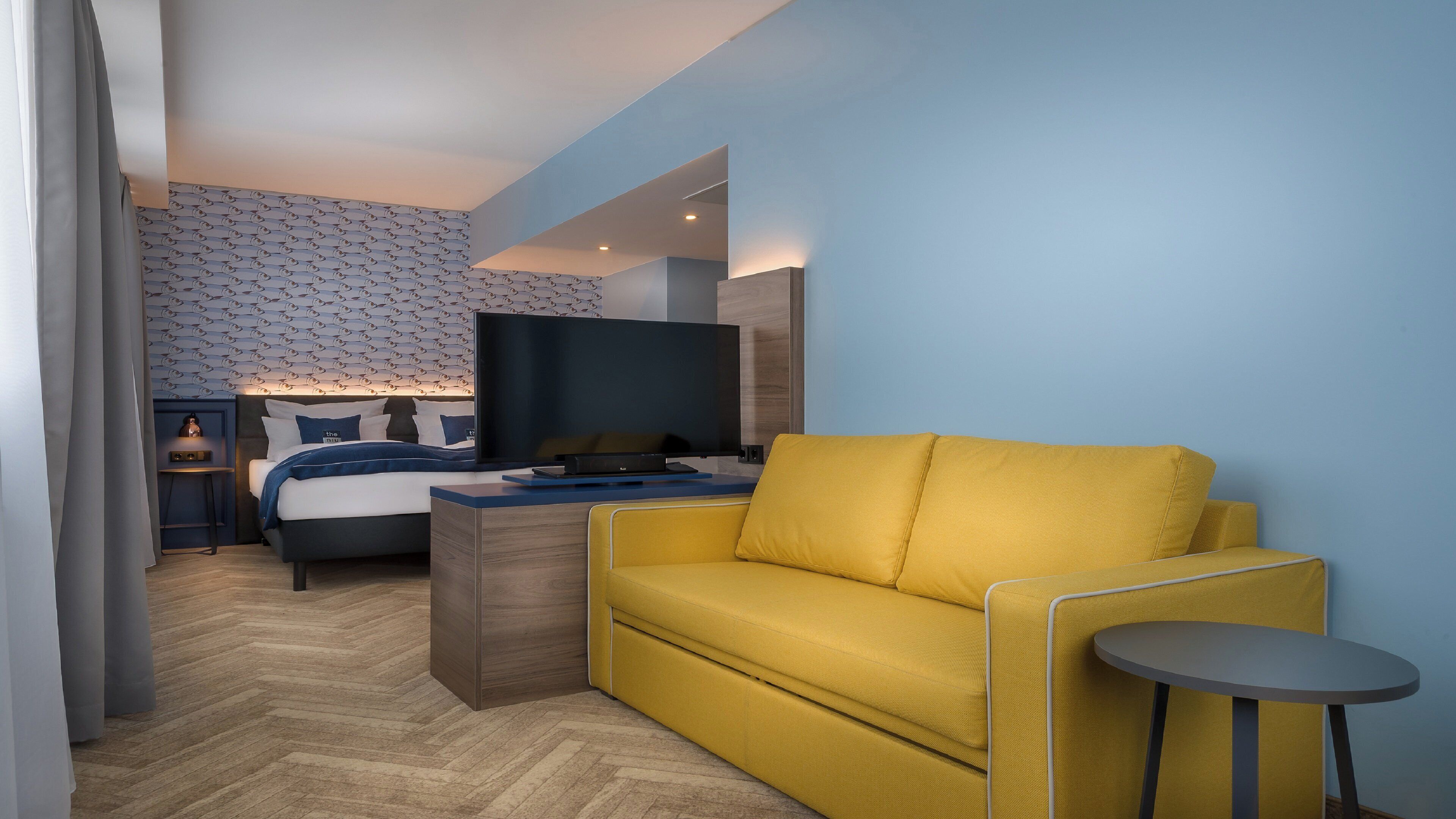 Une chambre d’hôtel avec un lit, une télévision, un canapé jaune, une petite table et un mur à motifs.