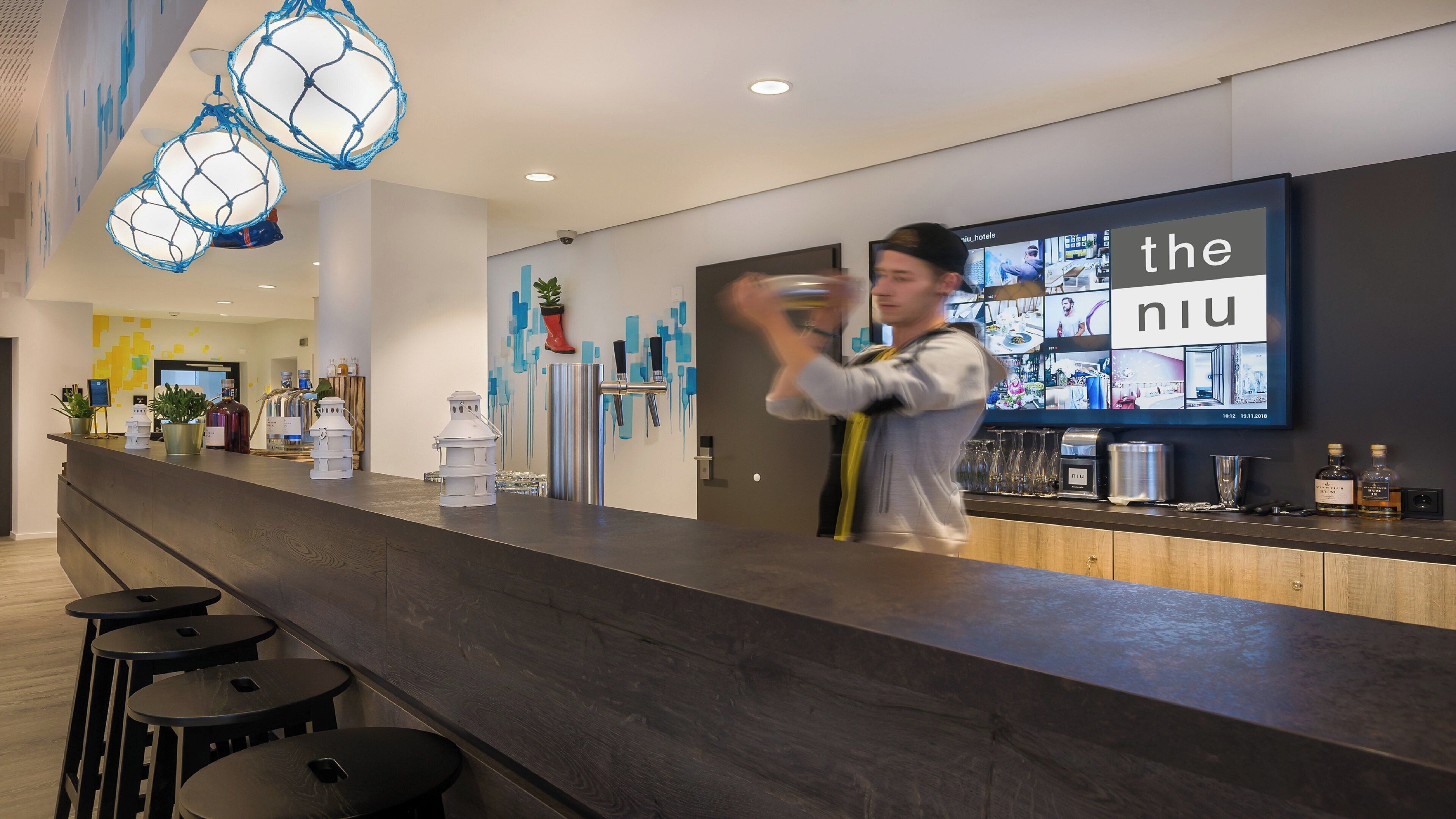 Una moderna area reception di un hotel, con un bancone in legno, sgabelli da bar e un grande schermo digitale che mostra il nome dell'hotel.