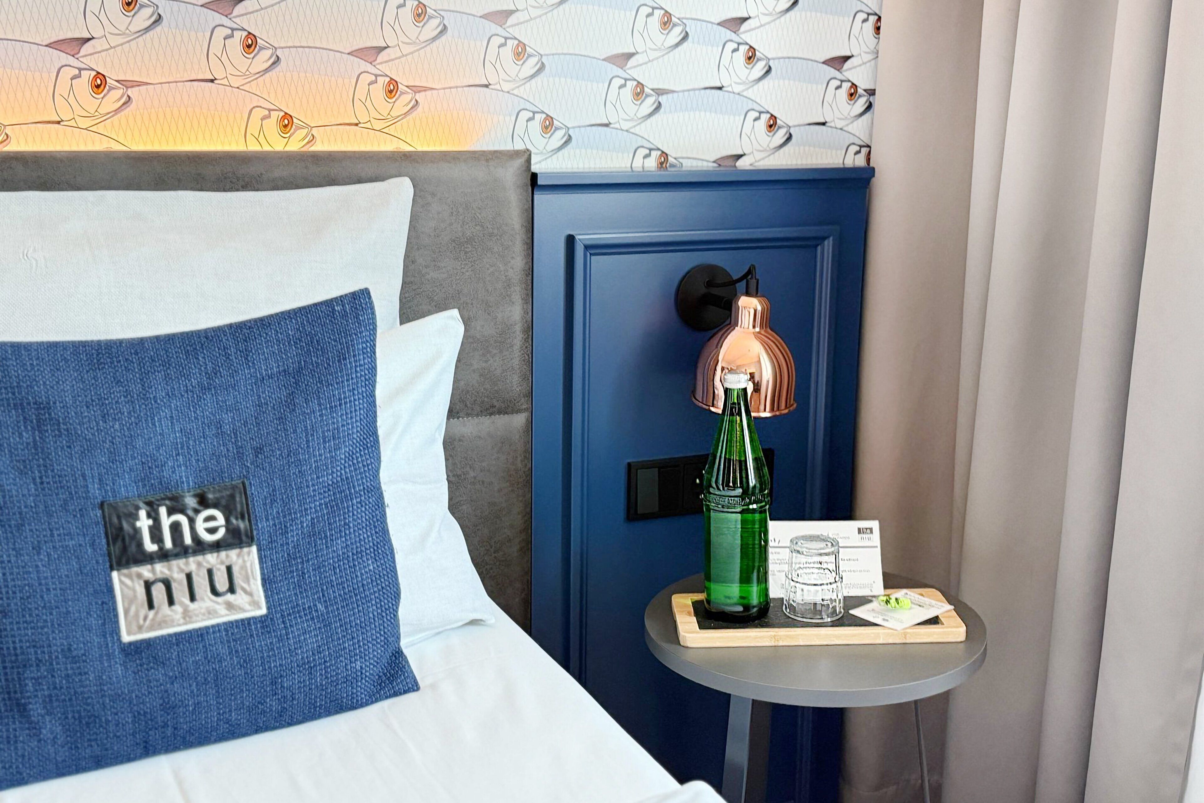 Ett hotellrum med en säng som har en kudde med logotypen för ”the nu”, ett sängbord med en grön flaska, en lampa och en meny.