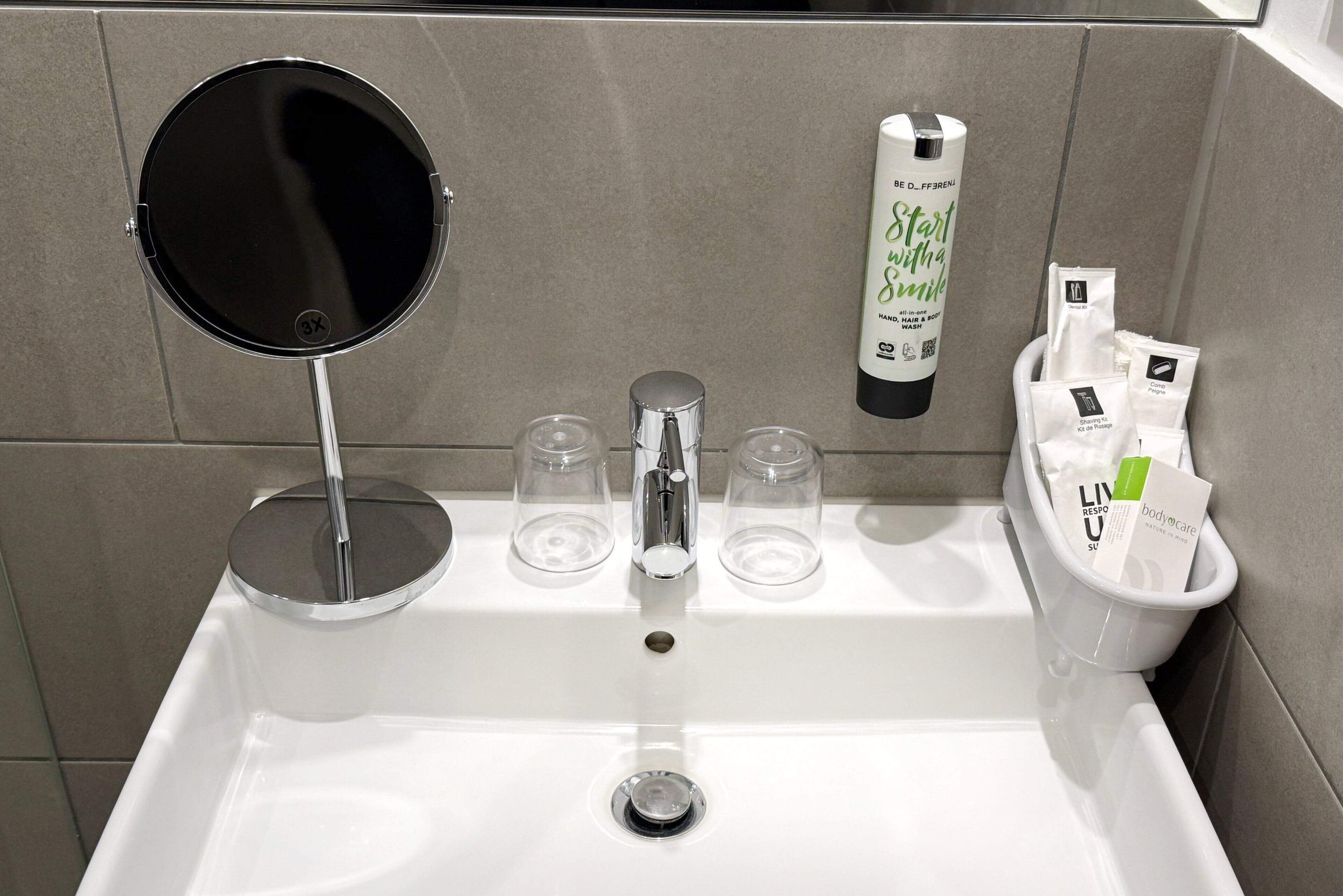 Een wastafel met een ronde spiegel, twee glazen bekers, een zeepdispenser en een bakje met toiletartikelen.