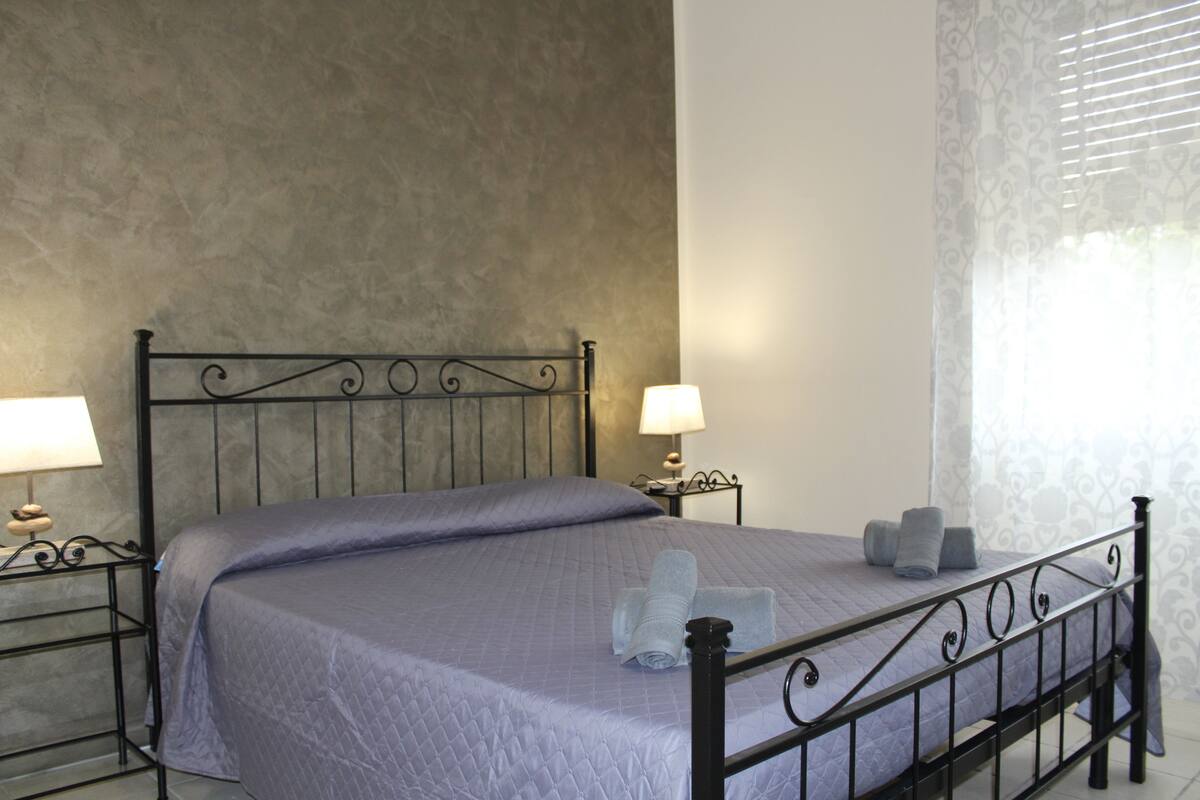 A neatly made bed with a quilted bedspread, two pillows, and rolled towels on a metal frame bed in a room with a textured wall and a window with curtains.