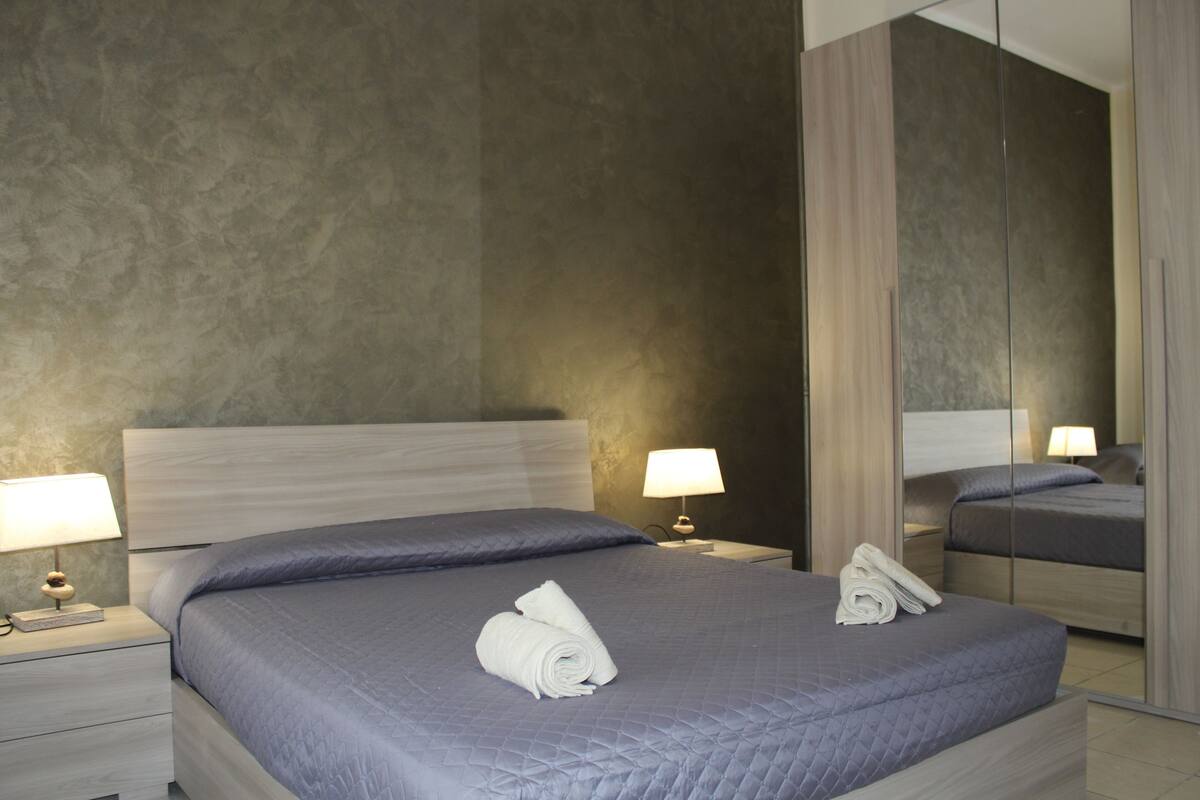A modern hotel room with a large bed, two bedside tables, a nightstand, a mirror, and a wall with a textured pattern.