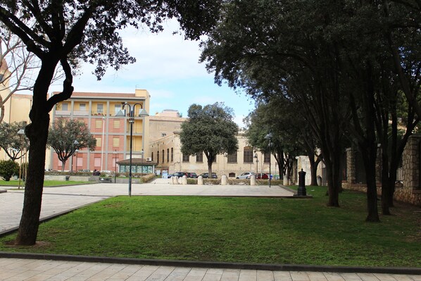 A park with a paved walkway, green grass, and trees. There are parked cars and a building with a yellow facade in the background. - Cagliari 4u (Cagliari)