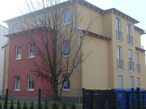 Un edificio de varios pisos con fachada roja y amarilla, un árbol en frente y un área cercada con contenedores azules.