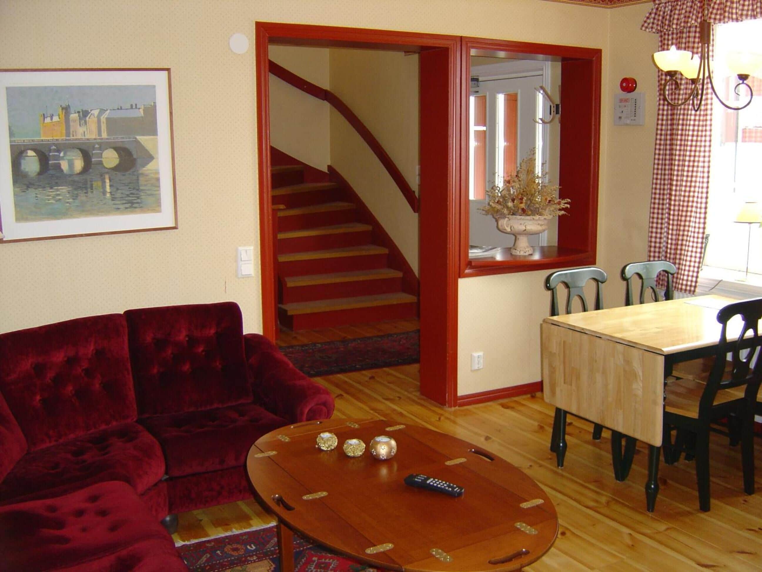 A living room with a red velvet sofa, a wooden coffee table with decorative items, a dining table with chairs, and a staircase leading to another room.