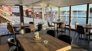 Een restaurant met houten tafels en stoelen, grote ramen met uitzicht op een besneeerd landschap en een rood gebouw.