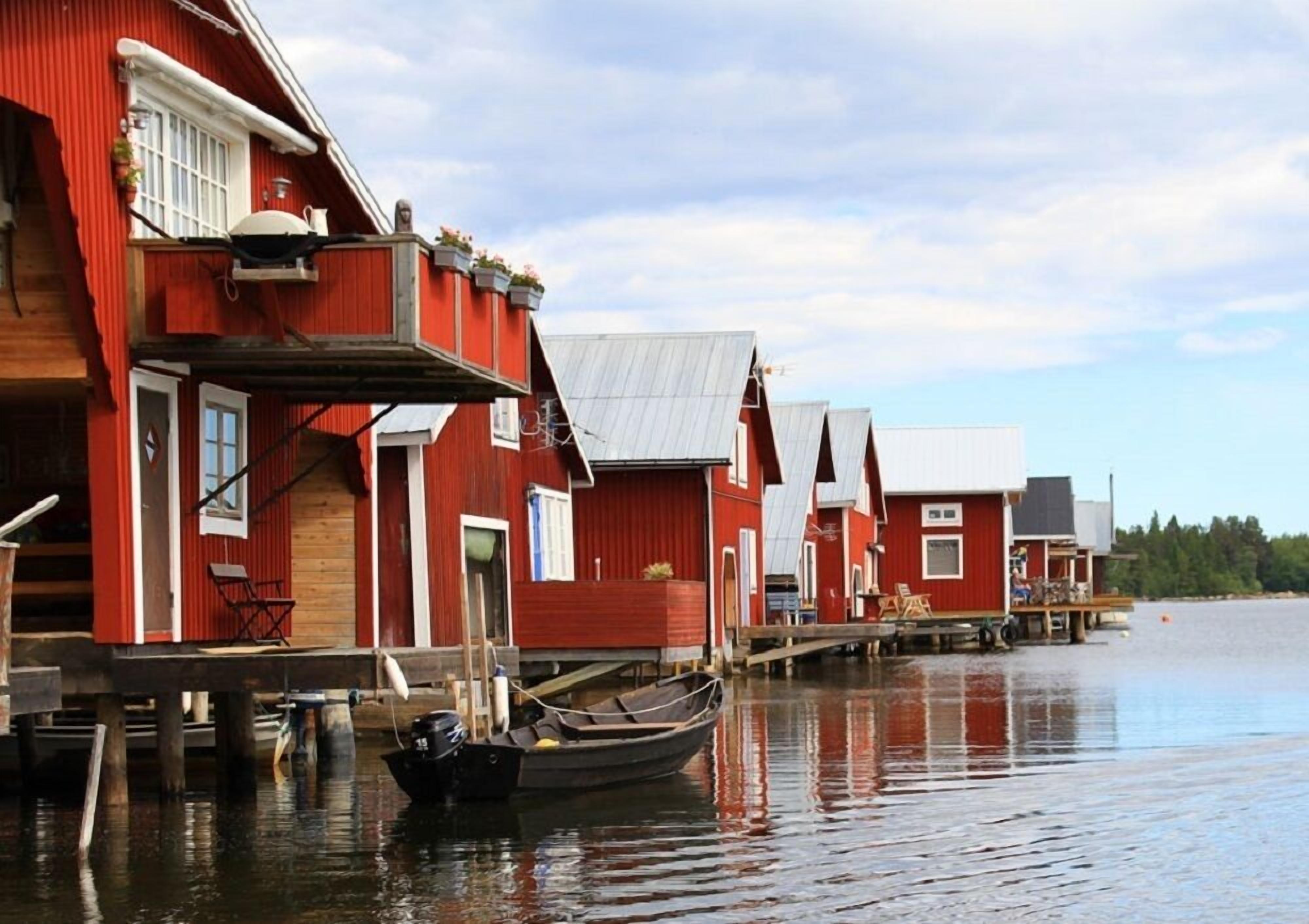 En rad röda trähus med en brygga som sträcker sig ut i stilla vatten, båtar förtöjda vid bryggan och en klar himmel ovanför.
