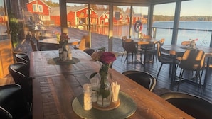 Una terraza de madera con mesas y sillas, con vista a un cuerpo de agua y edificios rojos al fondo.