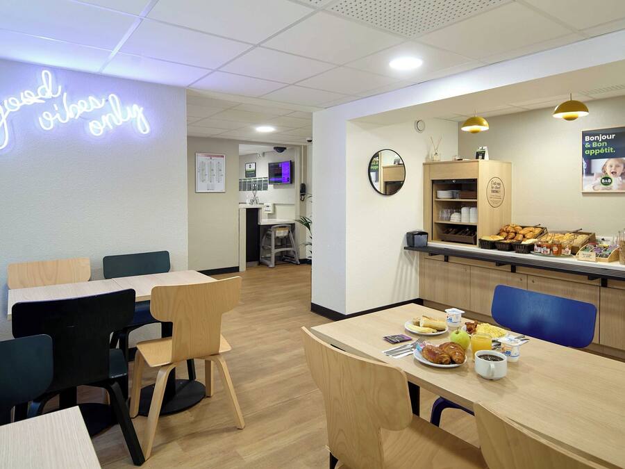 Un café moderne avec des tables et des chaises en bois, un panneau lumineux au néon, un espace cuisine et une variété de plats proposés pour le petit-déjeuner, disposés sur le comptoir.