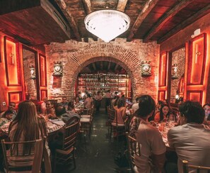 Een restaurant met bakstenen muren, houten balken en een kroonluchter. Tafels zijn gedekt met stoelen, en mensen dineren.