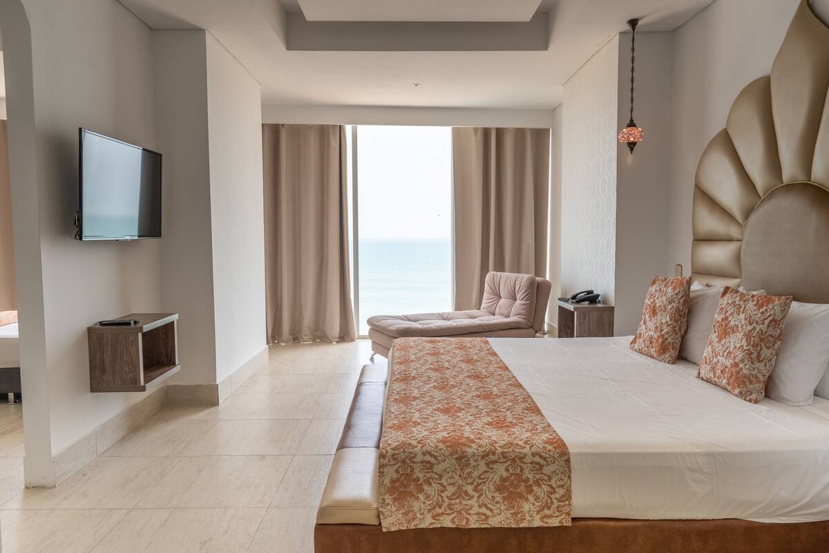 Une chambre d’hôtel moderne dotée d’un grand lit, d’un téléviseur à écran plat fixé au mur, d’un coin salon avec un fauteuil et d’une vue sur la mer grâce à de grandes fenêtres.