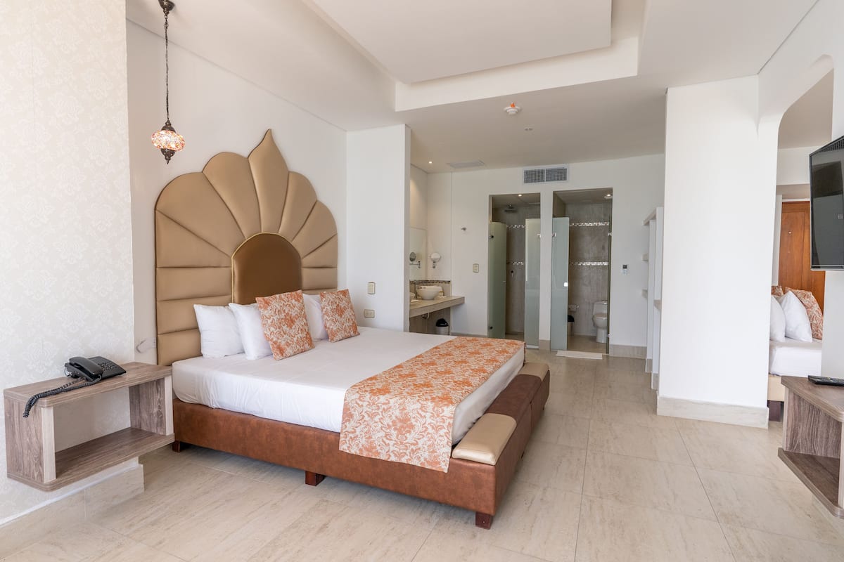 Une chambre moderne avec un grand lit, des tables de chevet, une télévision et une salle de bain visible à travers une porte ouverte.
