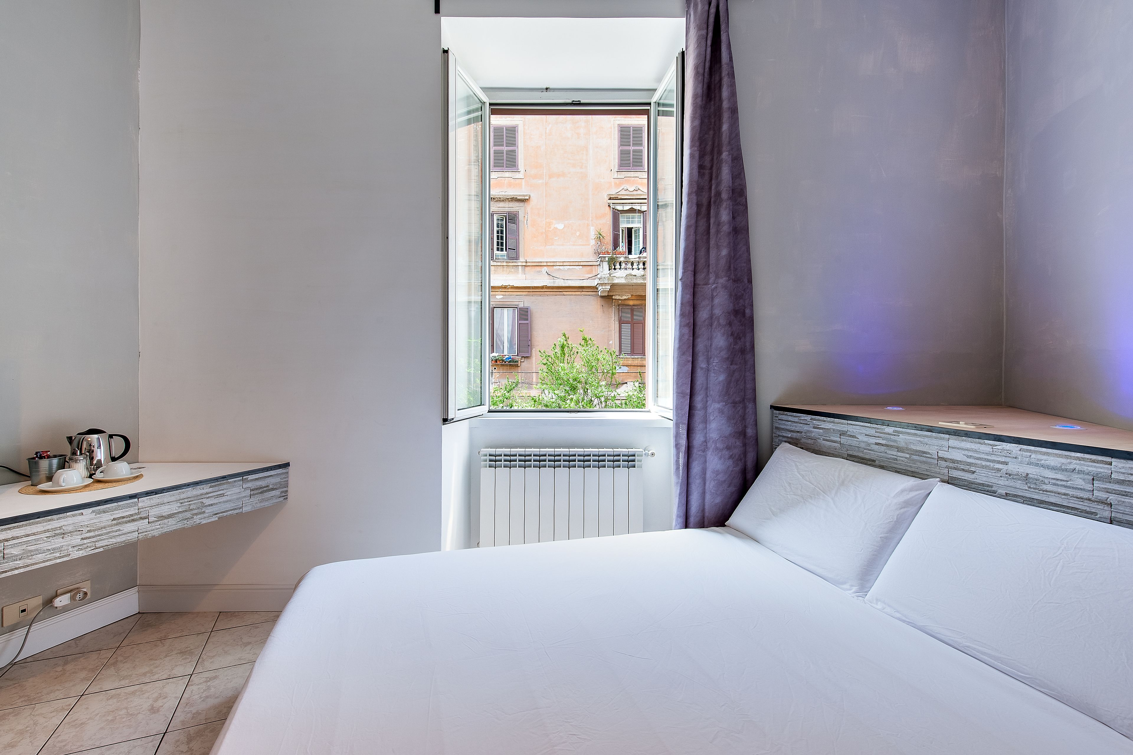 A hotel room with a bed, a window with a view of buildings, a bedside table with a kettle, and a radiator.