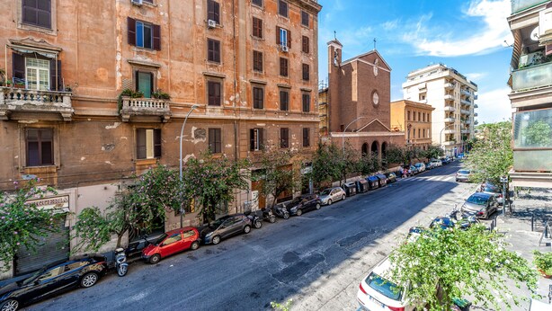 Vista de la calle con una fila de coches aparcados, un edificio con varias ventanas y balcones, y un cielo azul despejado.