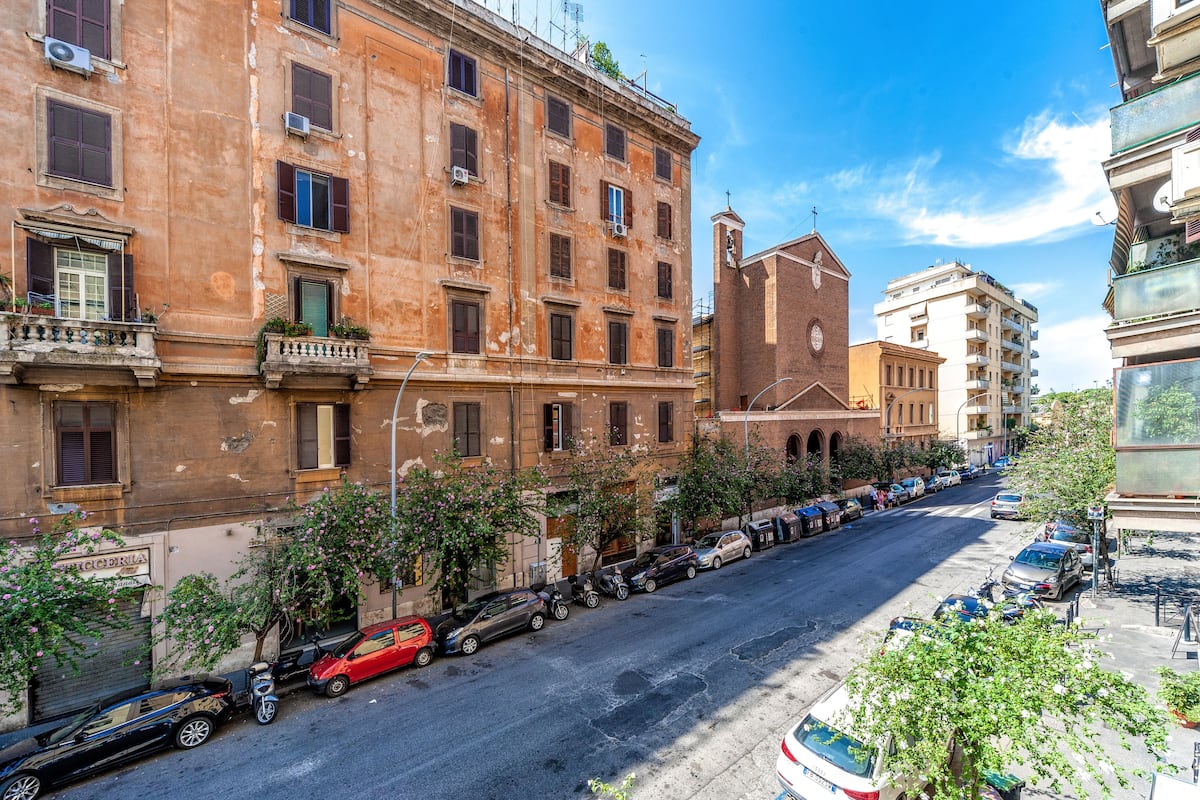 A street view with a row of parked cars, a building with multiple windows and balconies, and a clear blue sky.