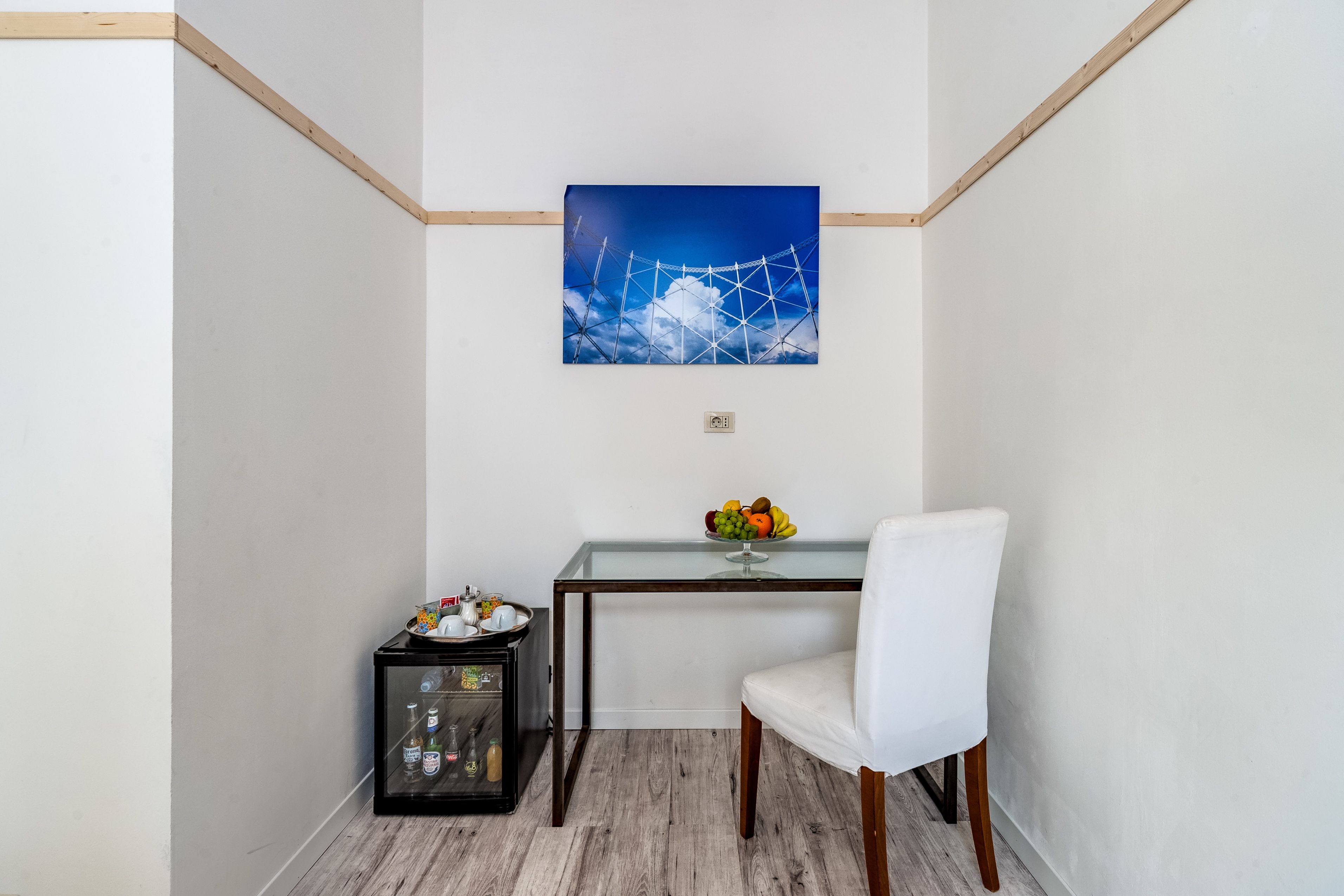 A corner room with a white chair, a small table with a fruit bowl, and a wall-mounted picture of a bridge.