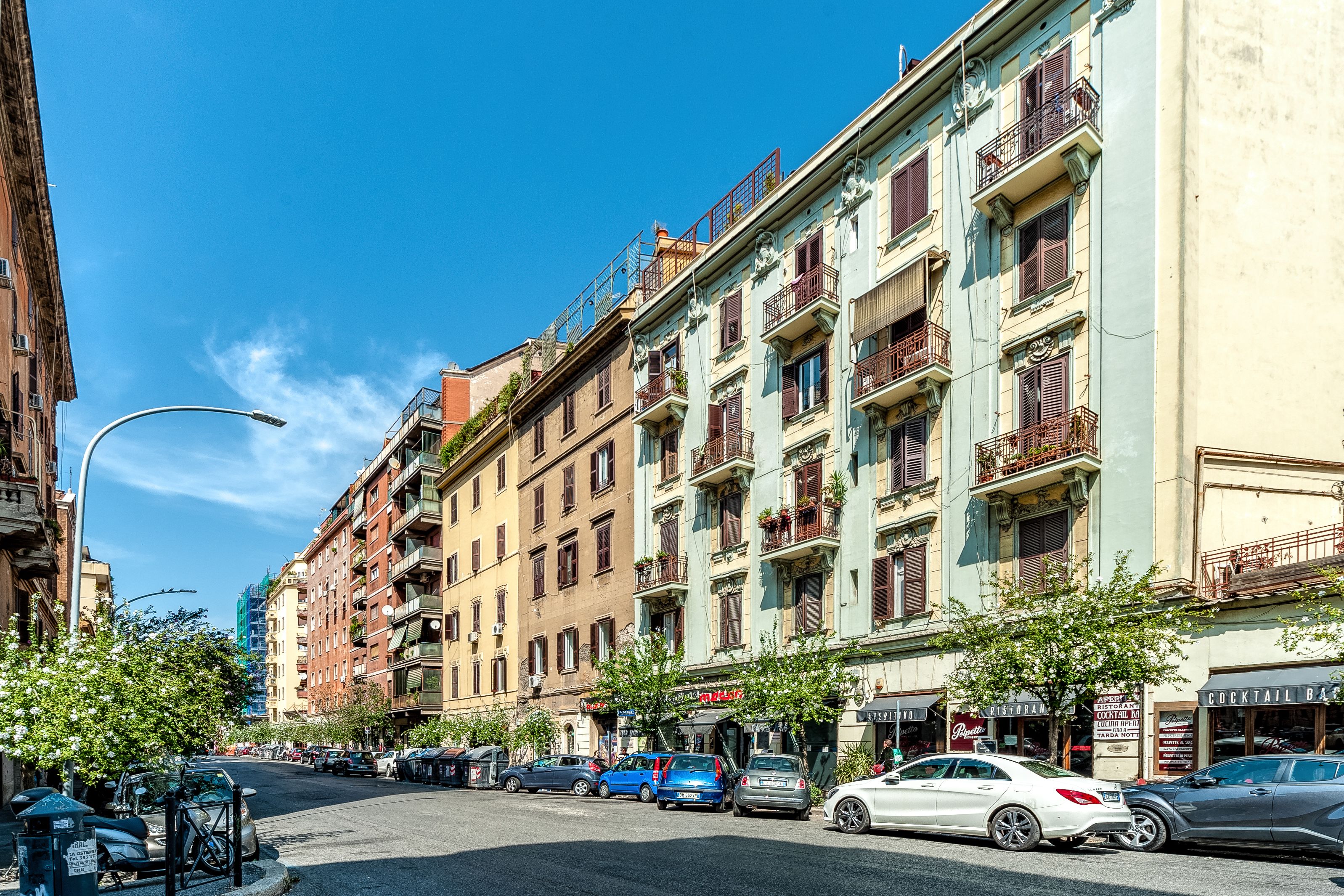 A row of multi-story buildings with balconies, parked cars, and a clear blue sky.