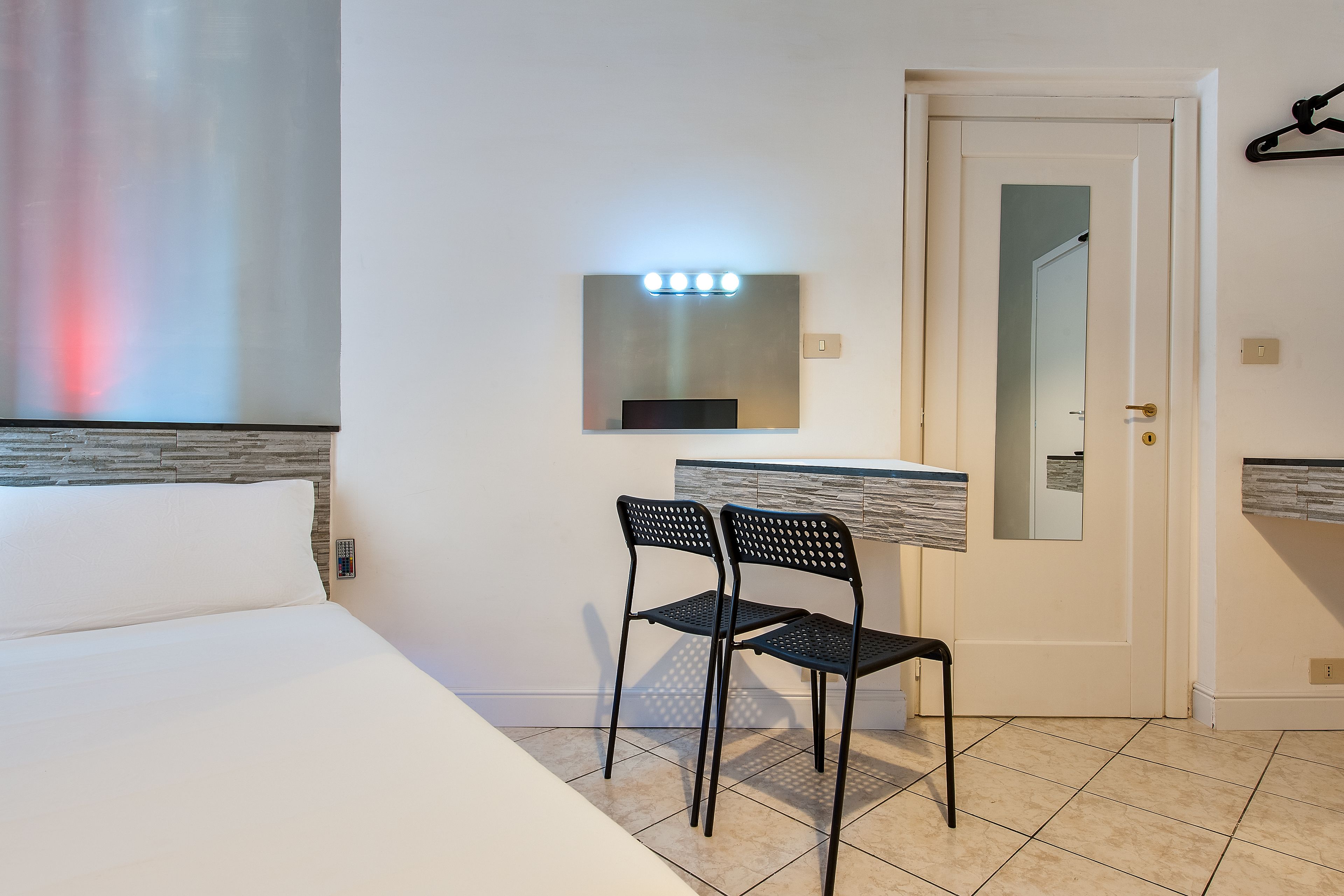 A hotel room with a bed, a desk with a mirror, two chairs, and a wall-mounted light fixture.