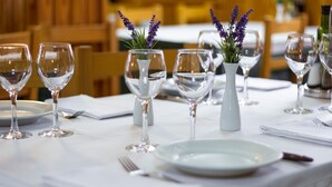 Una mesa lista para comer, con vasos transparentes y platos blancos, acompañada de pequeños jarrones con flores moradas.