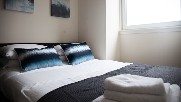 A neatly made bed with a white and gray blanket, two pillows with a blue and white pattern, and a folded white towel on the side. - Suite Apartments Aberdeen (Aberdeen)