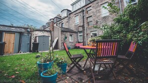 A stone building with a garden area featuring a table and chairs, potted plants, and a wooden bench. - Suite Apartments Aberdeen (Aberdeen)