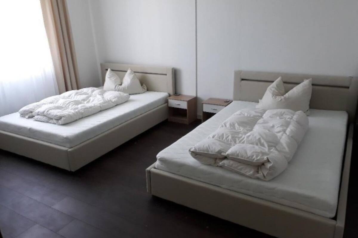 A hotel room with two single beds, each with a wooden headboard and white bedding. There are bedside tables with drawers and a small nightstand between the beds.