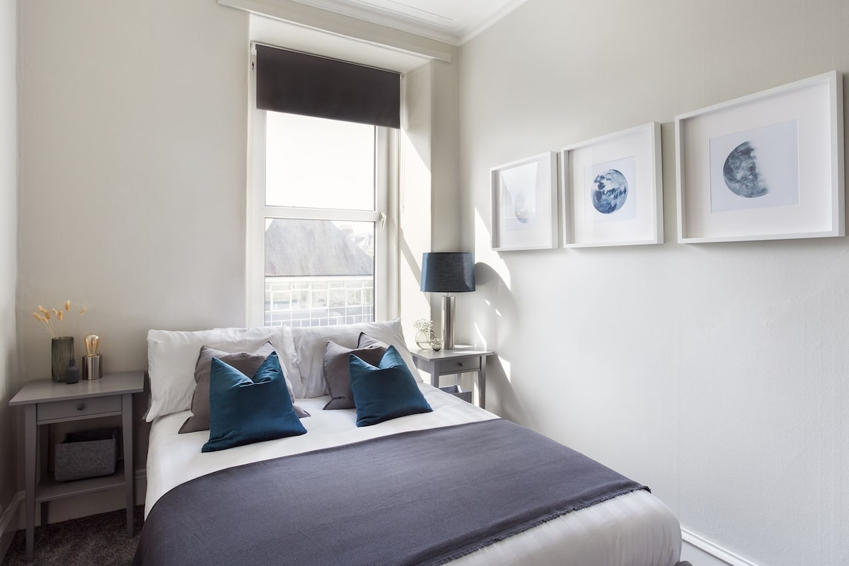 Ein Schlafzimmer mit Bett, Nachttischen, einem Fenster mit Jalousie und gerahmten Bildern an der Wand.