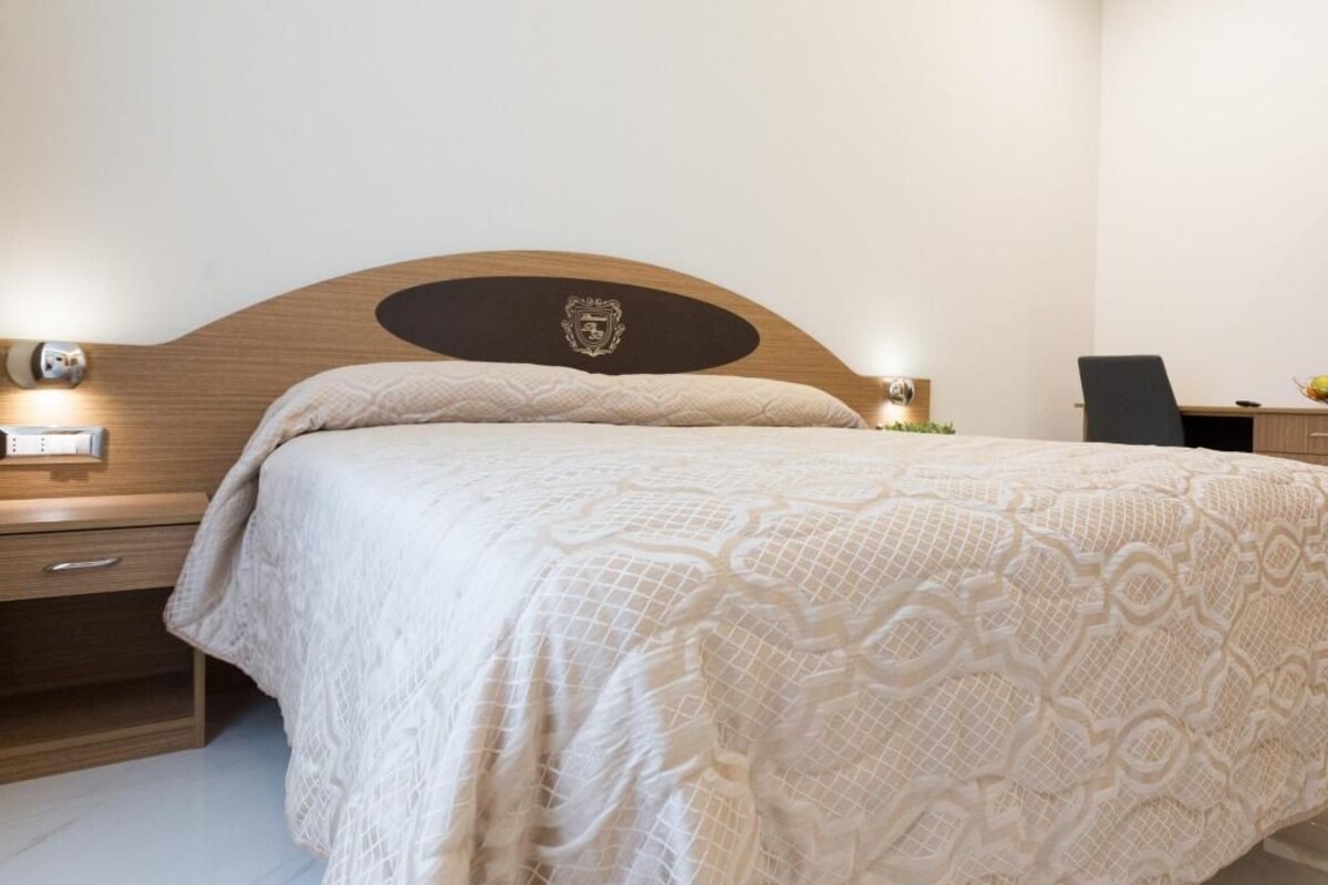 A bedroom with a wooden headboard, a bed with a patterned bedspread, bedside tables with lamps, and a small desk with a chair.