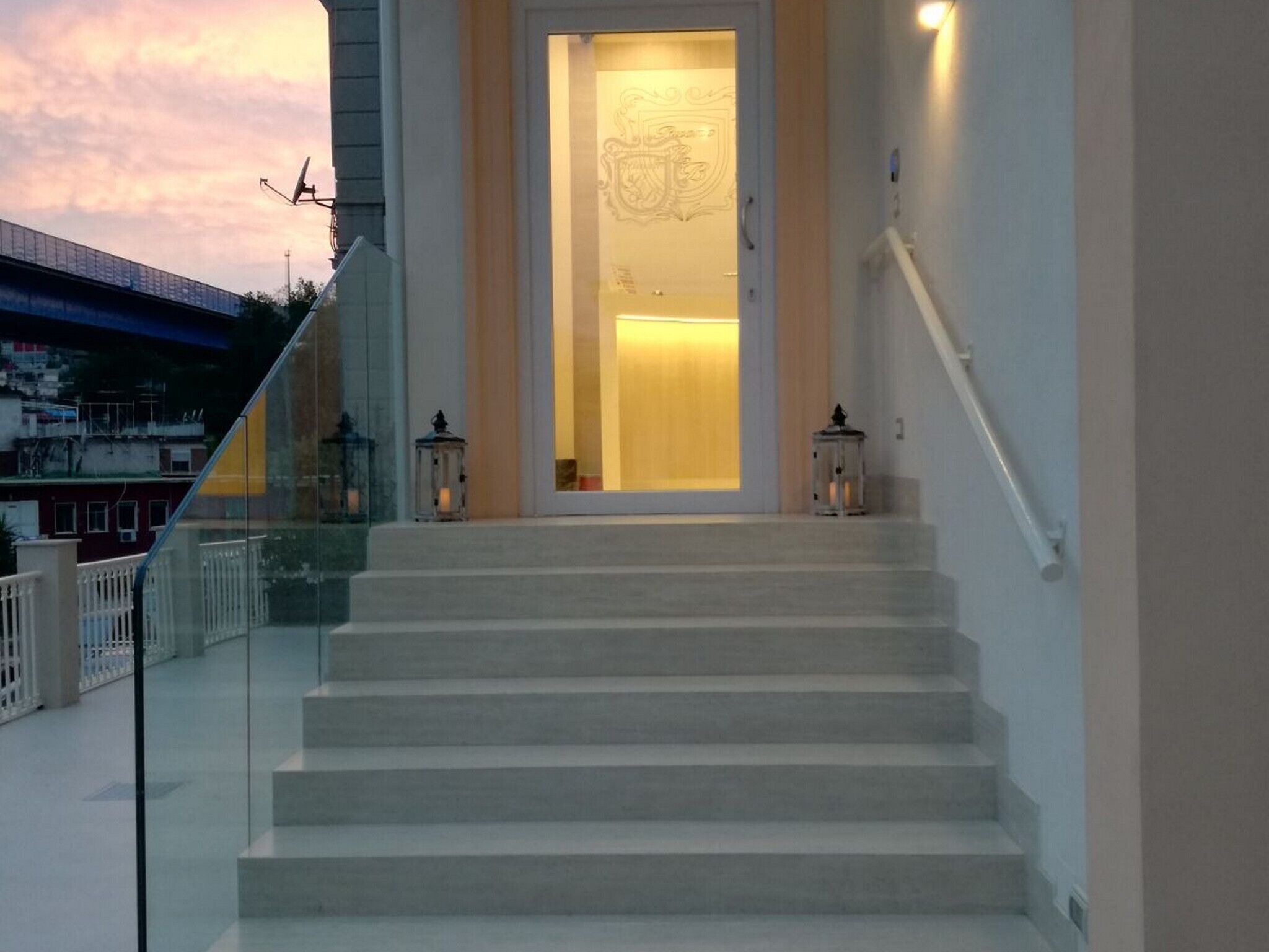 Uma escadaria leva a uma porta com painel de vidro, dois candeeiros e um corrimão.