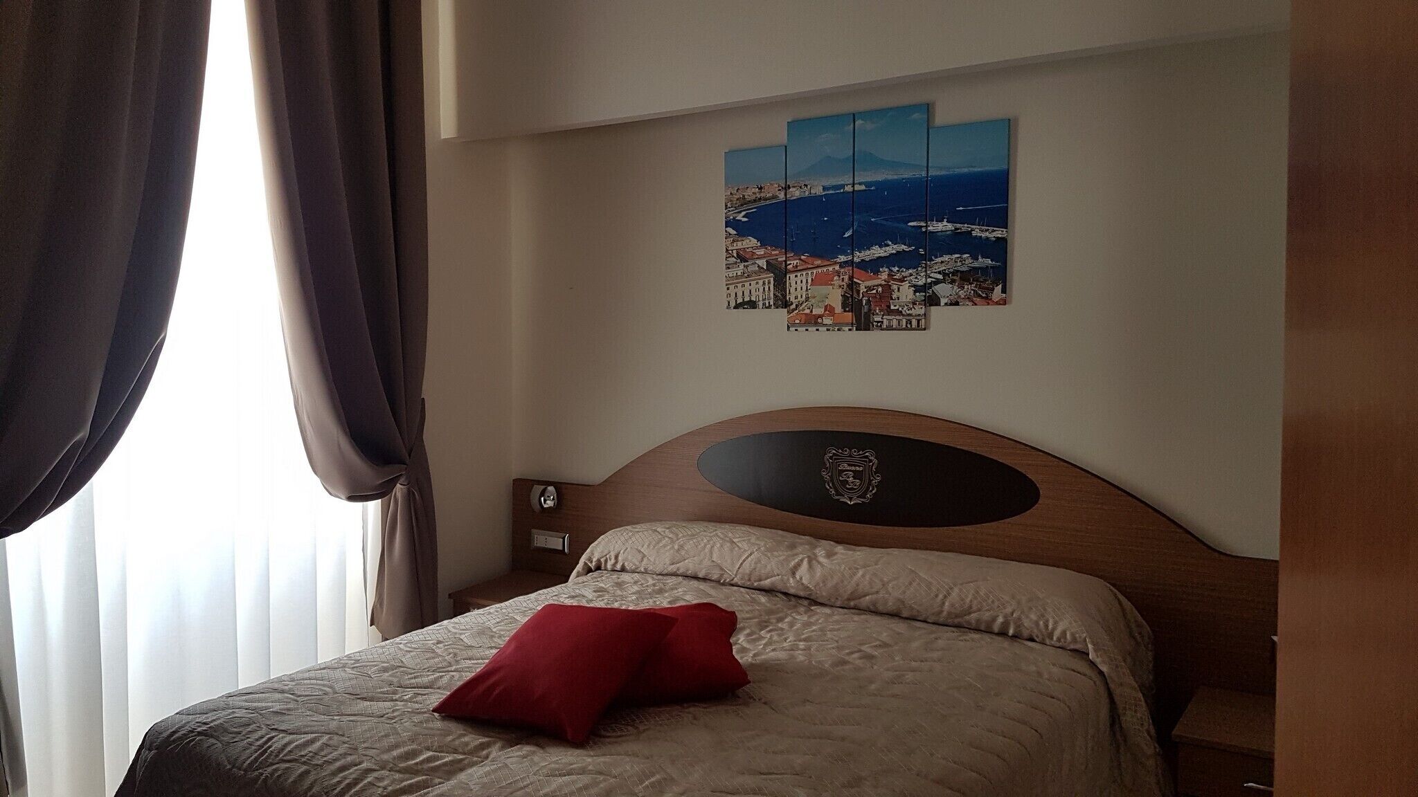 A hotel room with a bed, a red pillow, a wall-mounted artwork depicting a coastal scene, and a wooden headboard with a crest emblem.