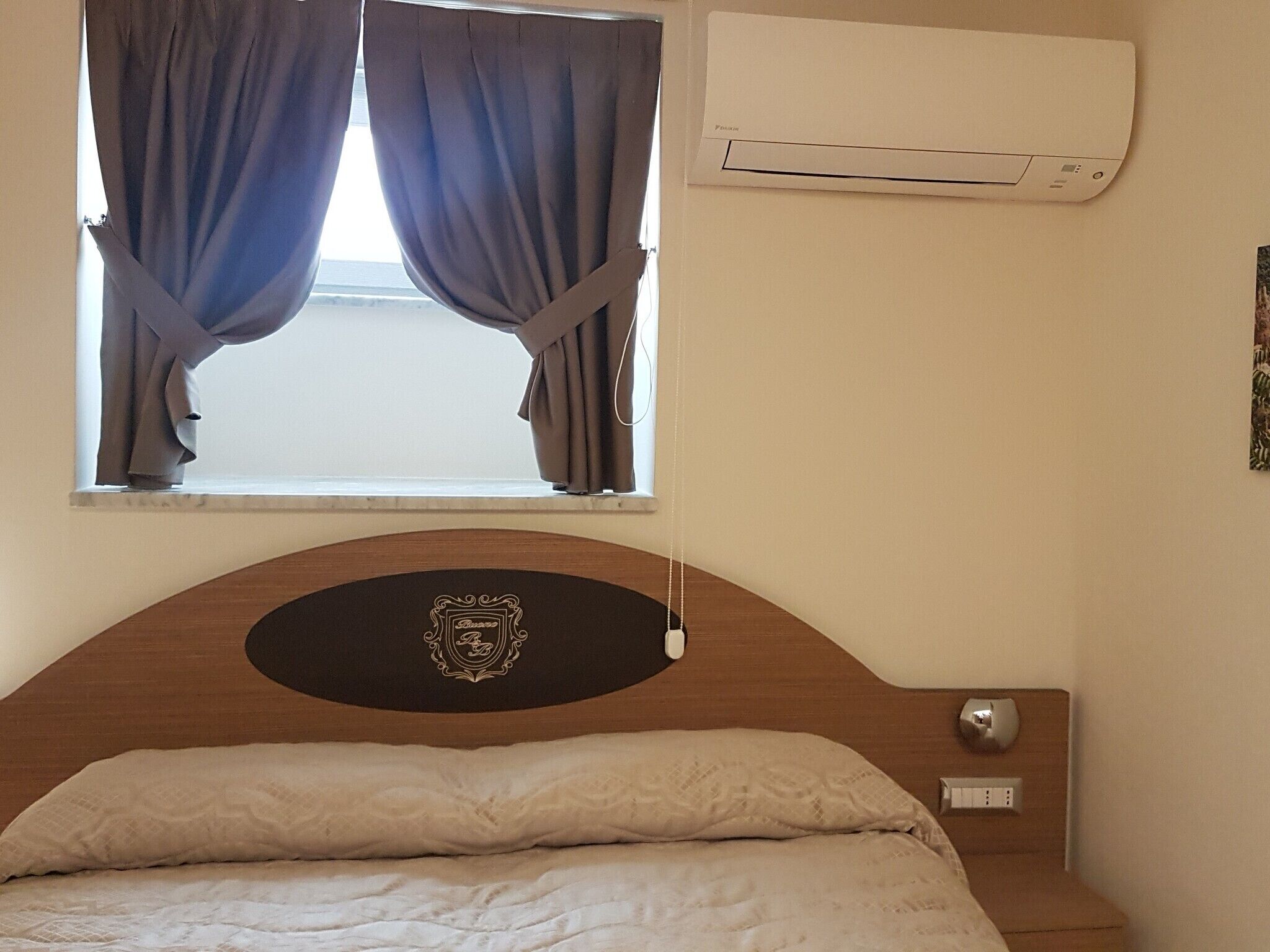Un dormitorio con cama, ventana con cortinas, equipo de aire acondicionado y un emblema decorativo en la cabecera.