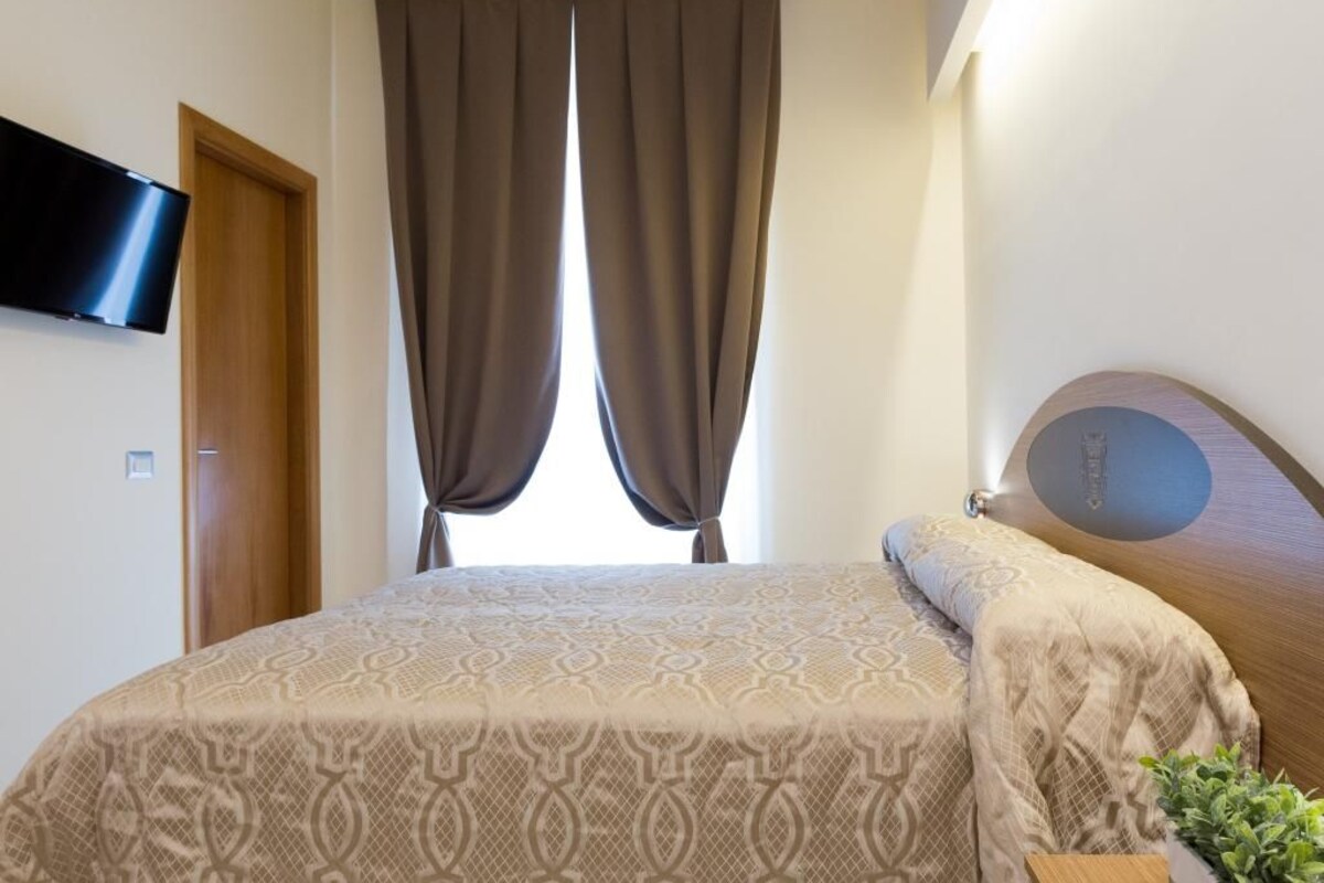 A bedroom with a bed, curtains, a television mounted on the wall, and a plant.