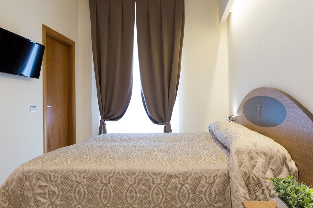 A bedroom with a bed, curtains, a television on the wall, and a plant.