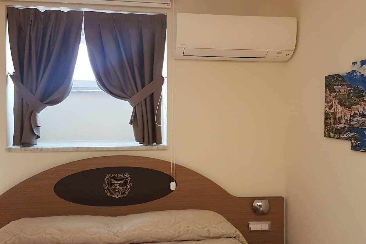 A hotel room with a bed, a window with curtains, an air conditioning unit, a wall-mounted light, and a partial view of a map.
