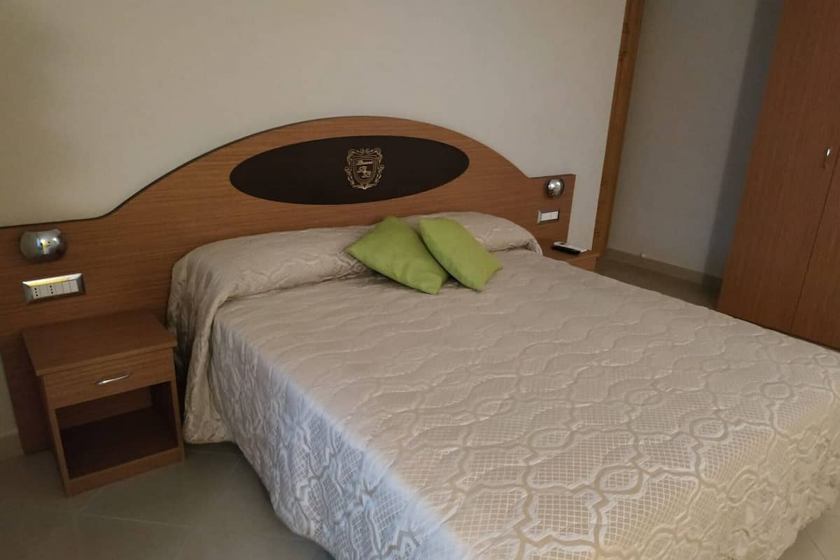 A neatly made bed with a patterned bedspread and two pillows, a wooden headboard with a crest emblem, a bedside table with a lamp, and a wooden wardrobe.